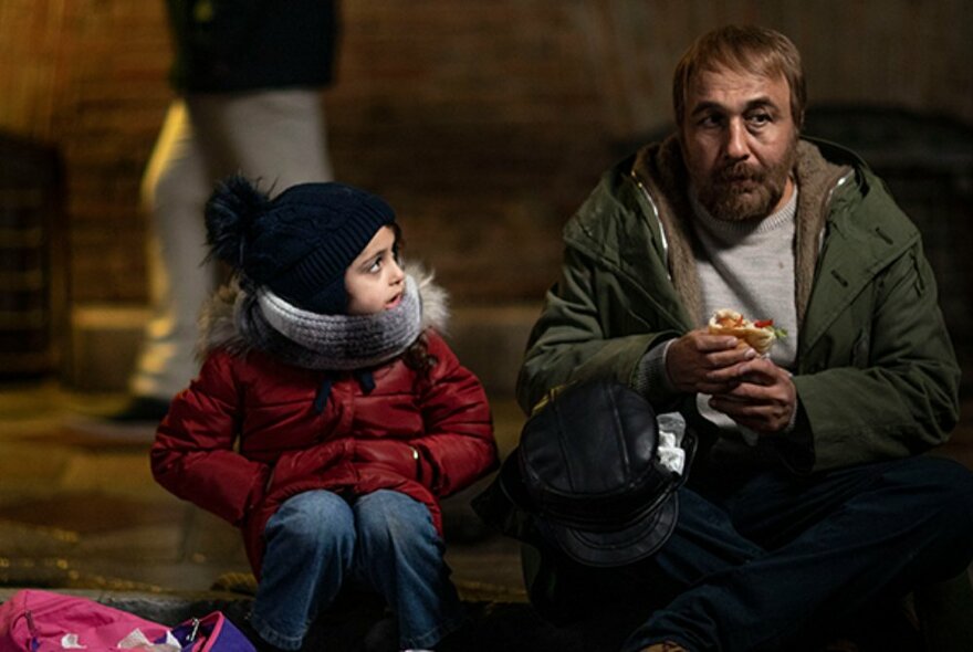 A still image from a film showing an adult man sitting in the street beside a child in a red coat and warm clothes. 
