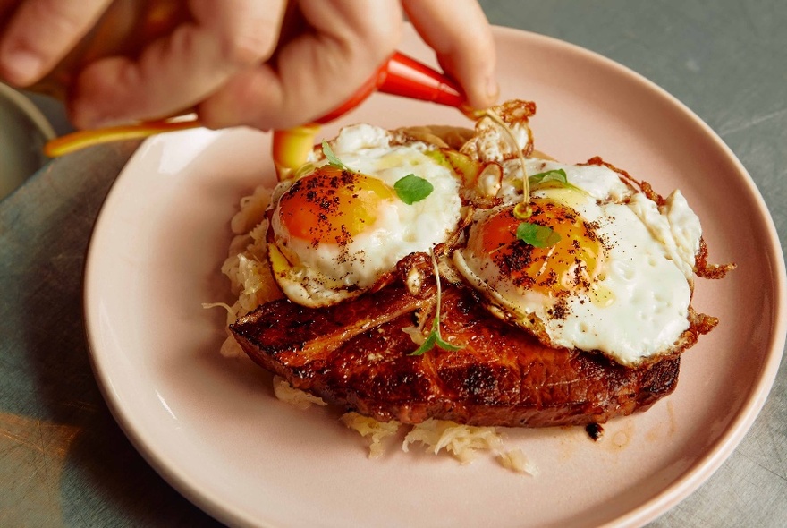 A hand drizzling olive oil onto a plate of fried eggs on top of a thick piece of bacon.