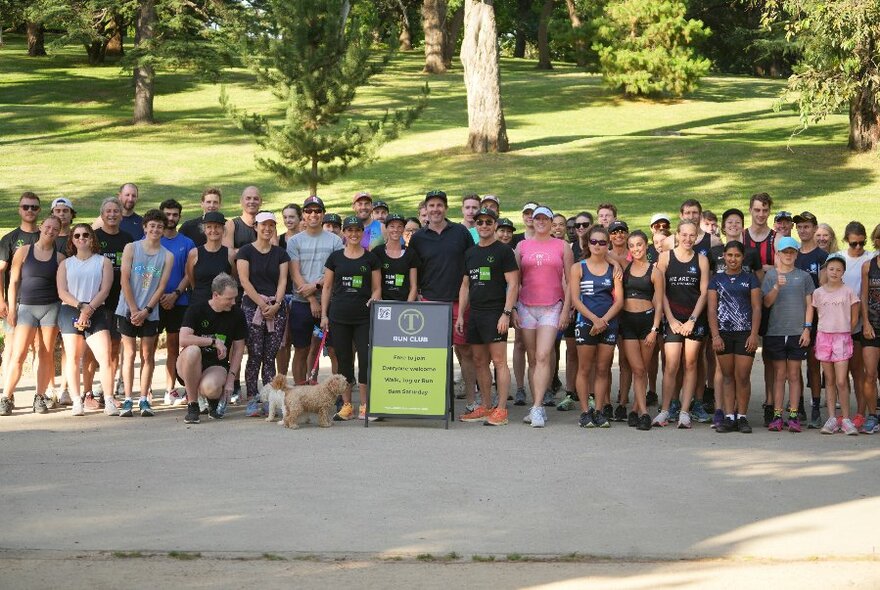A large group of people in active wear posing behind a sign in a park. 
