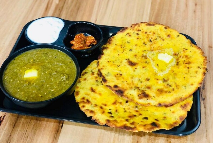 Sectioned black dish with naan bread, spinach curry and condiments.