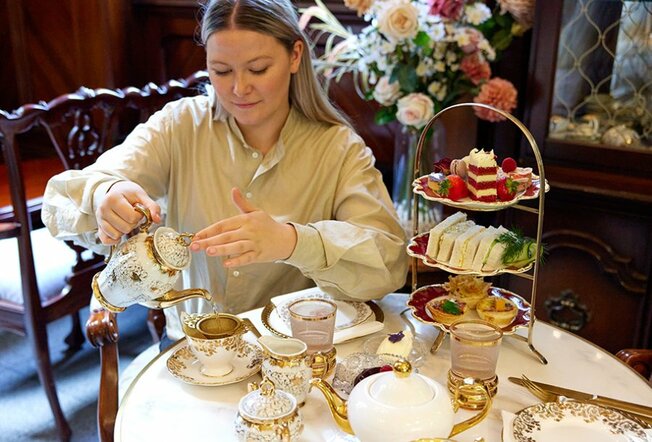 A woman pouring tea at a high tea with a tiered stand of food.