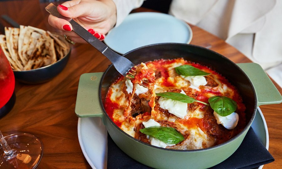 A forkful of lasagne from a large round dish, topped with mozzarella and fresh basil. 