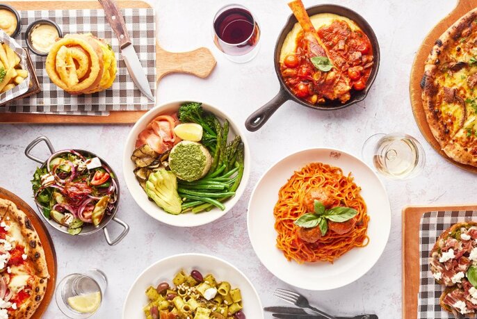 Overhead view of a table filled with dishes of food including bowls of pasta, a pizza, a burger and chips, plus salads.