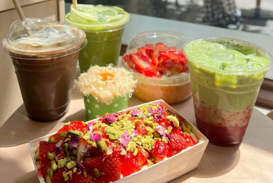 A selection of green drinks in plastic cups next to a strawberry-topped dessert in a takeaway container.