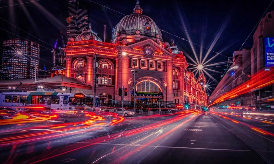 Flinders Street Station illuminated in red lights at night with blurred traffic passing. 