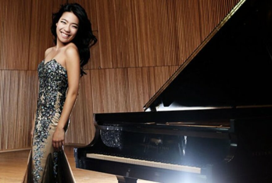 A smiling woman, the pianist Joyce Yang, wearing a sparkling strapless evening gown standing next to a grand piano on a stage.