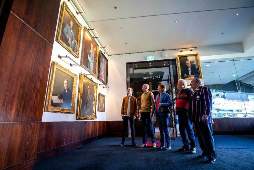 People on a tour looking at formal portraits on a wall in room with wood tones and blue carpet.