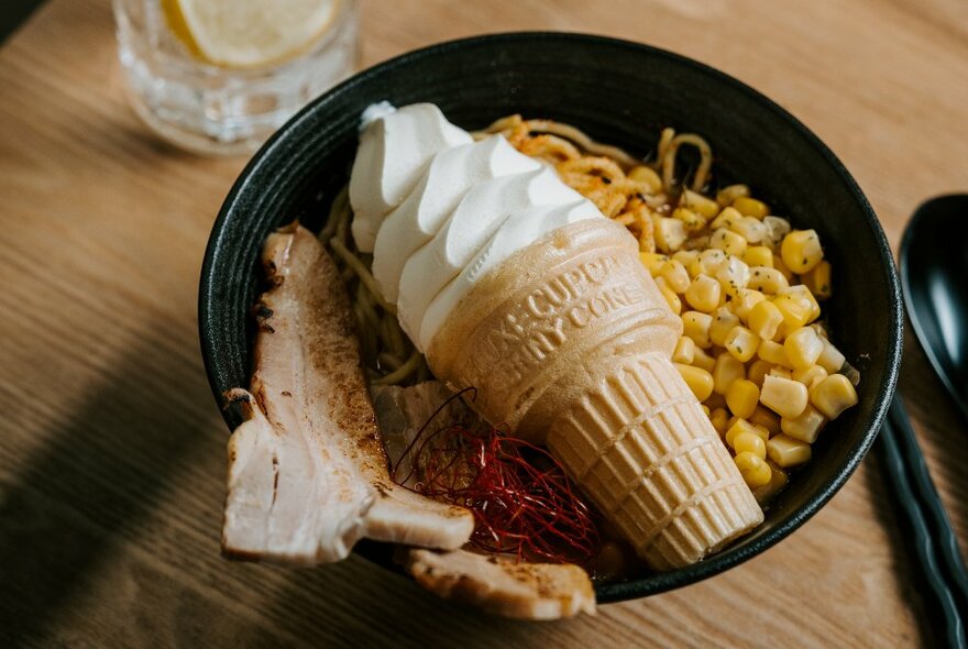 A bowl of ramen, pork, and sweetcorn kernels, with a soft serve ice cream resting on top.