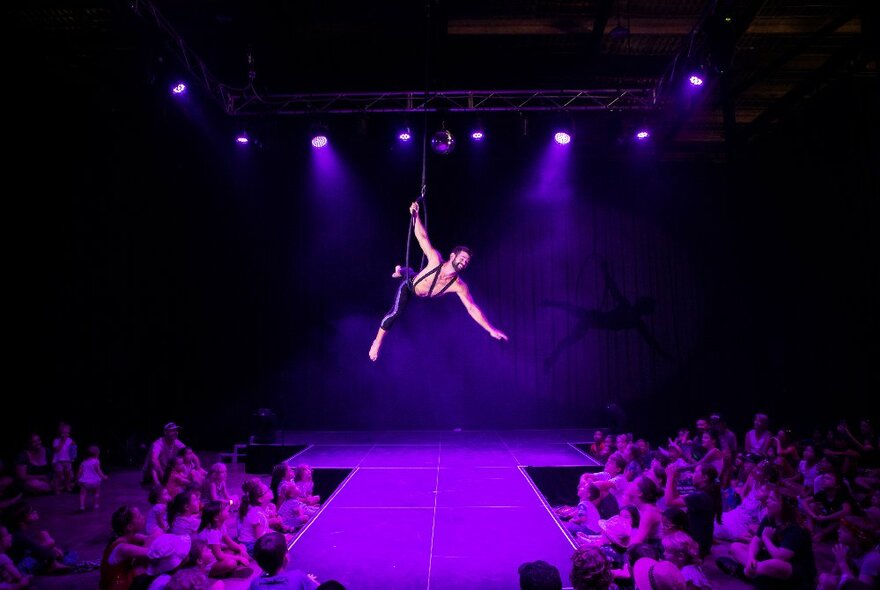 A circus performer wearing black tights flying above a darkened stage in a ring apparatus.