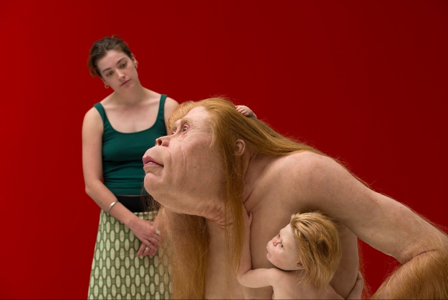 Person looking at an orangutan-person with child sculpture in a red gallery space