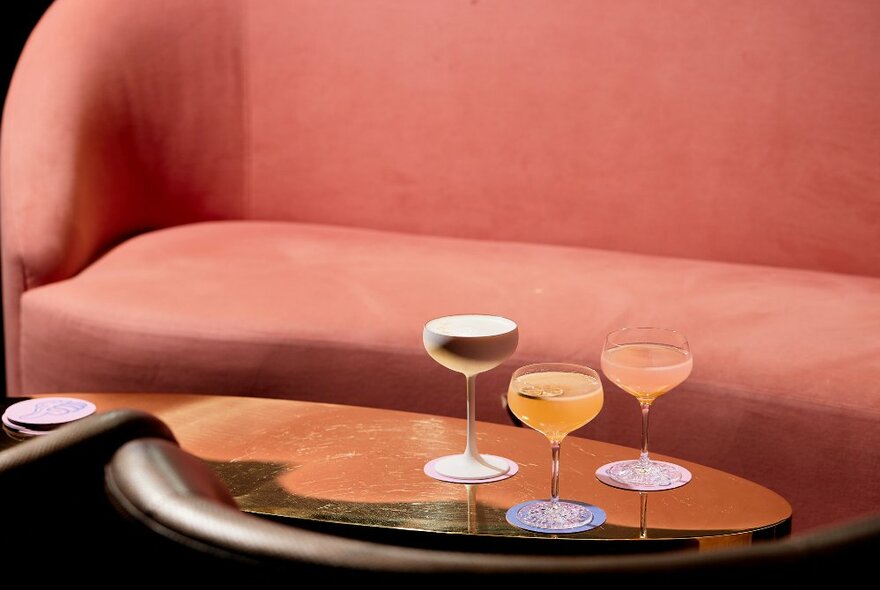 A salmon pink velvet couch with a table bearing three different cocktails. 