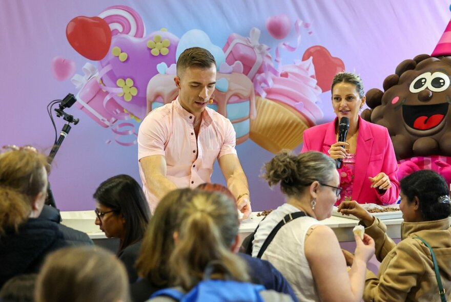 A person performing a cooking demonstration with a woman in a pink blazer holding a microphone, an audience at tables in the foreground. 