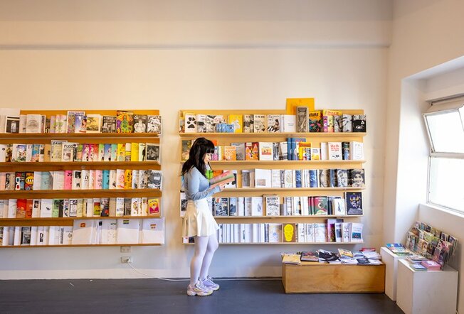 A woman reading a zine in a store with yellow shelves filled with zines.