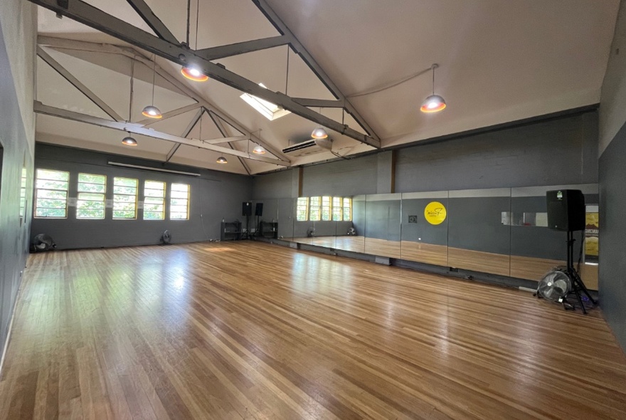 Large empty dance studio with a polished wooden floor, mirror along one wall, windows and high ceilings.