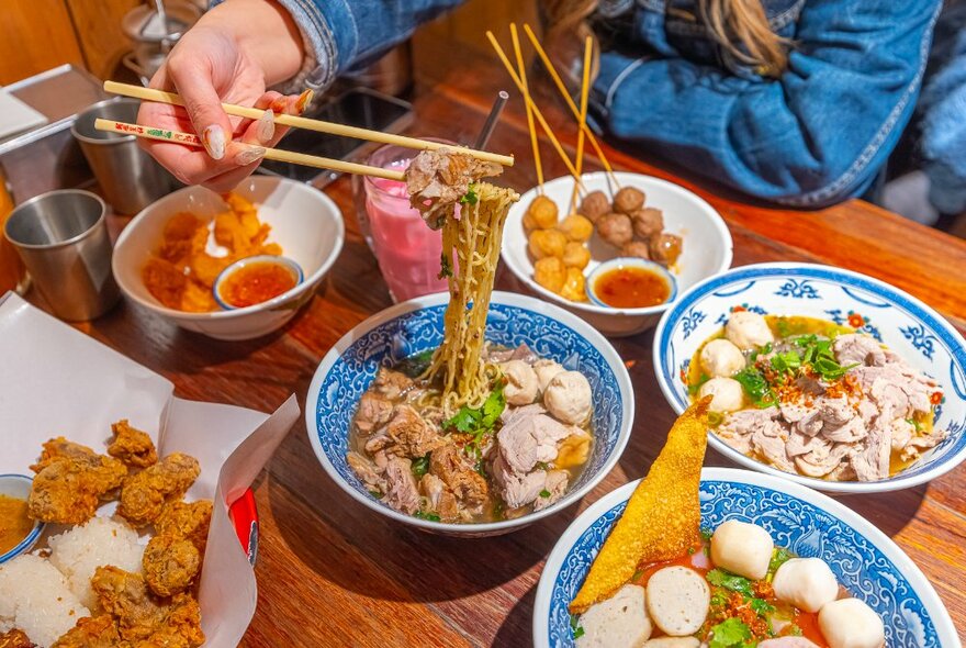 Person using chopsticks to pick up noodles from a soup bowl, other plates of food on the table including fried skewers. 