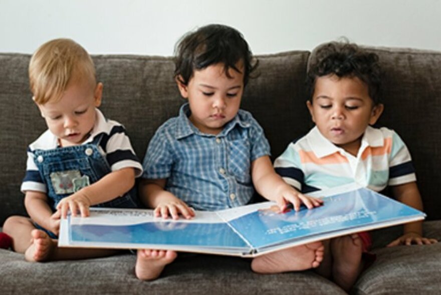 Three toddlers seated on a couch in a row, holding an open picture book on their legs.