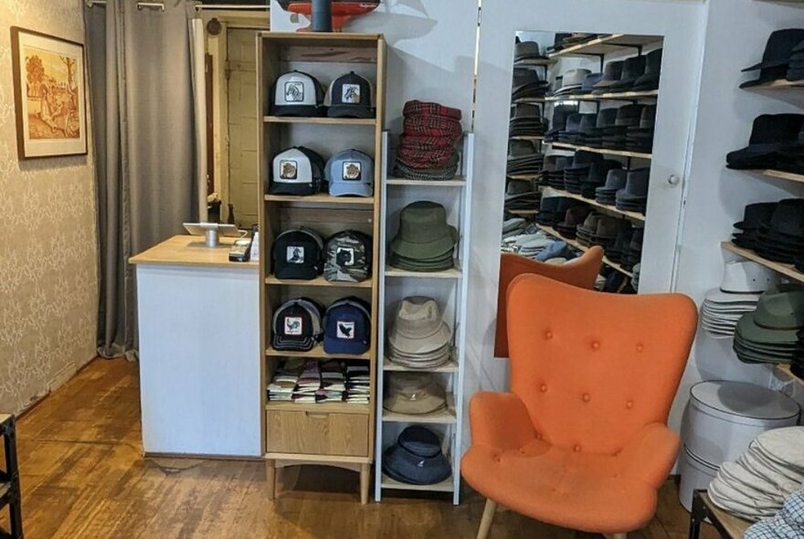Interior of a shop selling hats, with hats on shelves along a wall, a mirror, an orange armchair and a small counter service area.