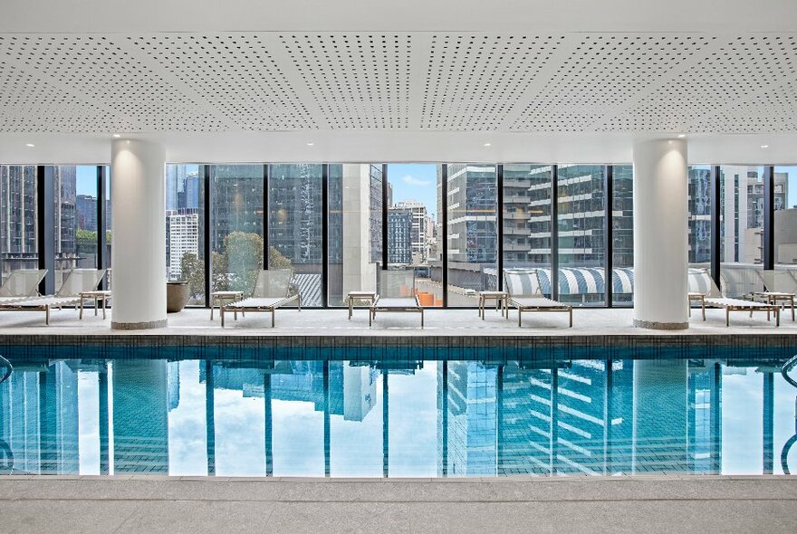 Large hotel indoor swimming pool with white pillars and views of city buildings.