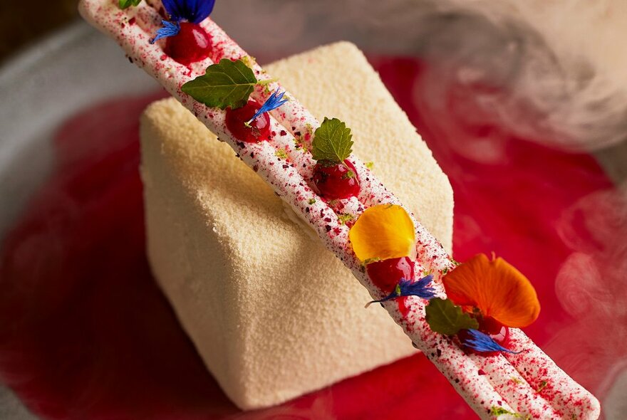 A modern looking dessert featuring a white cube and a specked garnish on a red plate. 