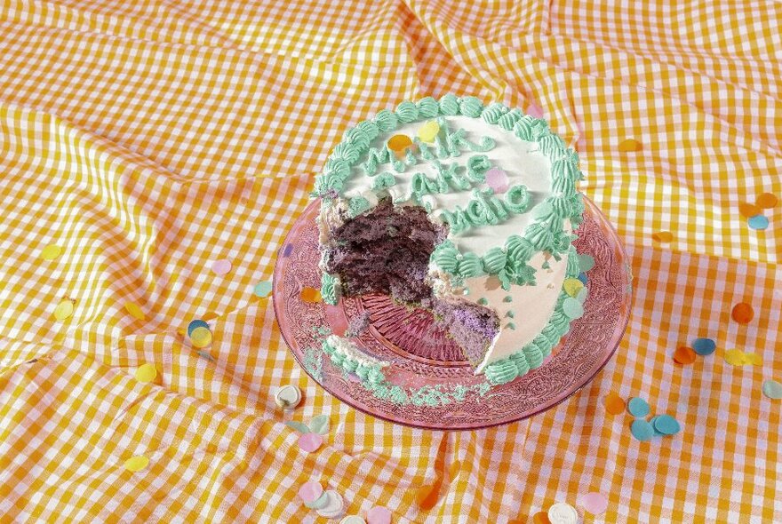 Looking down at a roughly chewed birthday cake on a yellow gingham tablecloth. 