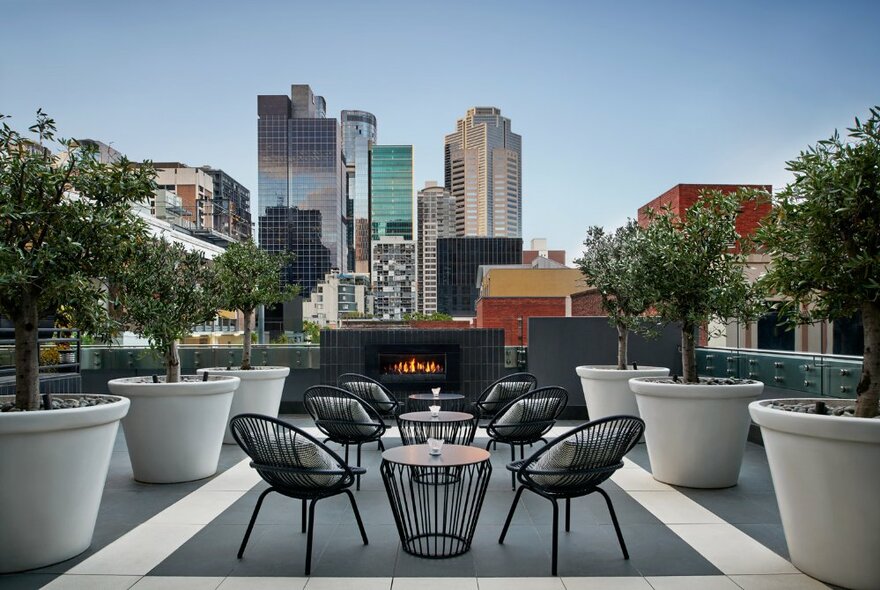 Hotel rooftop with trees in oversized pots, tables and chairs with fire pit overlooking city buildings.