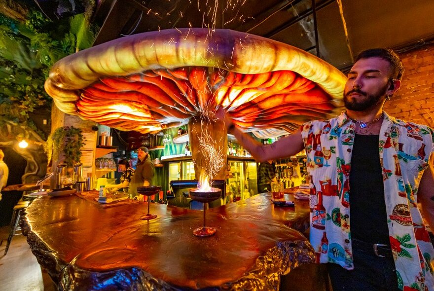 A bartender making a flaming cocktail in a themed bar under a giant mushroom.