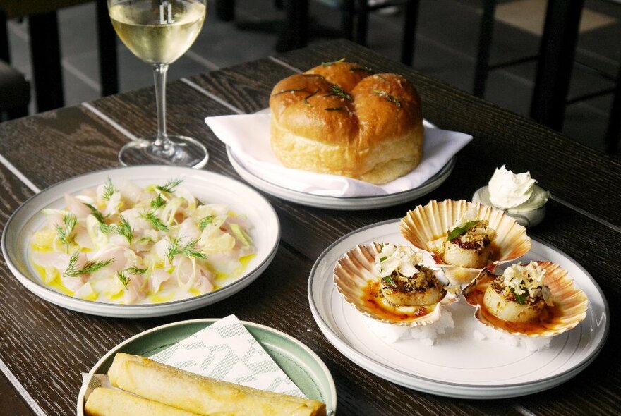 Several plates of food on a dining table including scallops in their shells, filled pasta, a small loaf of freshly baked bread and a glass of wine.