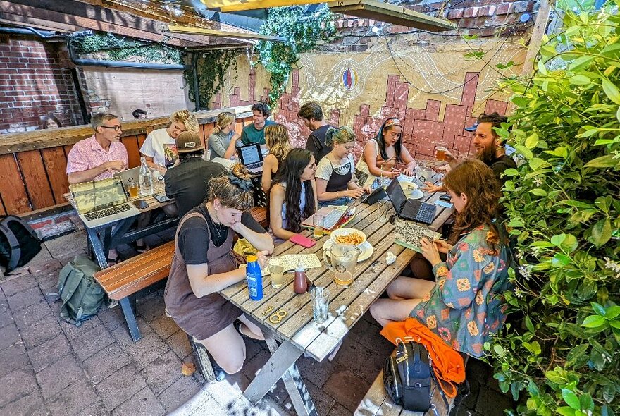 Group of people sitting across two tables in an outdoor beer garden, writing on laptops or in notebooks and drinking.