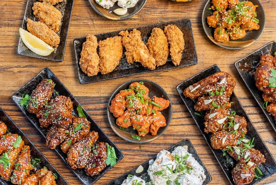 Overhead view of a table loaded with black dishes of food including crumbed meat, glazed meat and dips.