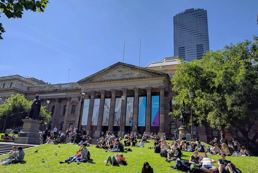 People seated on the lawn in front of the neo-classical columns and pediment of State Library Victoria.