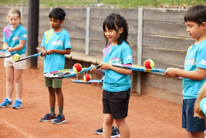 Four children in matching blue t-shirts practise balancing tennis balls on their racquets. 