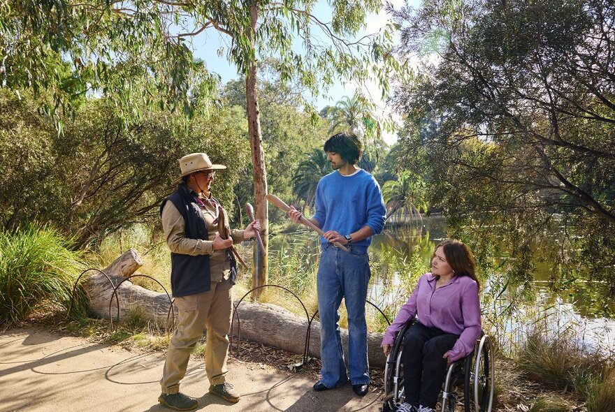 A guide in outdoor clothing chatting to two people in a garden. They are holding Aboriginal artefacts, the woman is using a wheelchair.