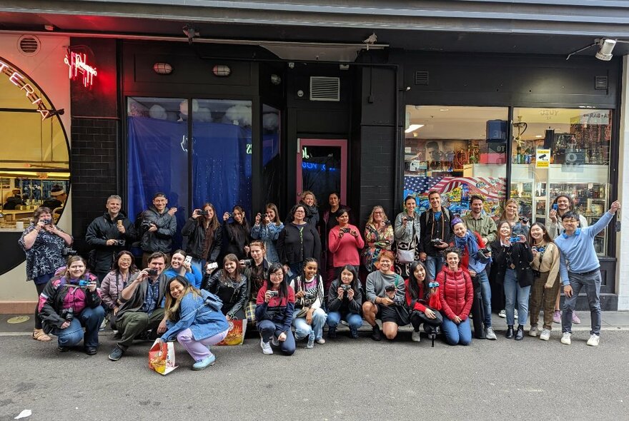 A group photo of a photography walking tour group with their cameras by a shopfront in a Melbourne laneway.