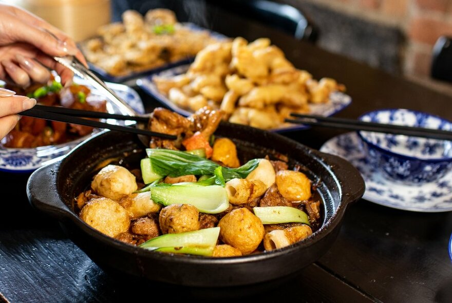 Close-up of black dish of food, others behind, hands with chopsticks on top.