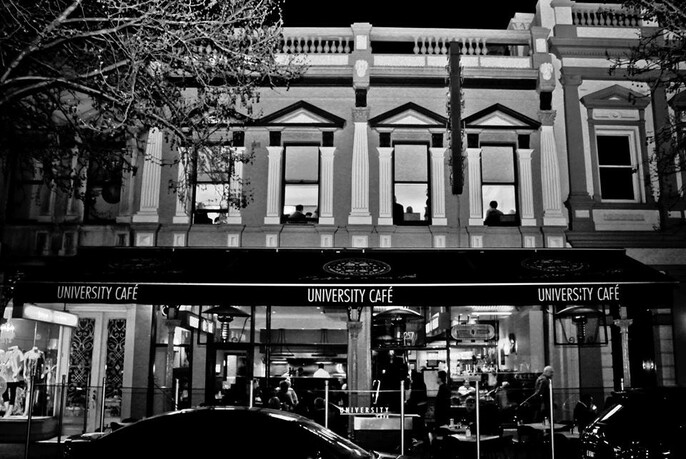 Cafe exterior at night with uplit windows with classical pediments on 1st floor and cafe windows, passing cars and signage at ground level.