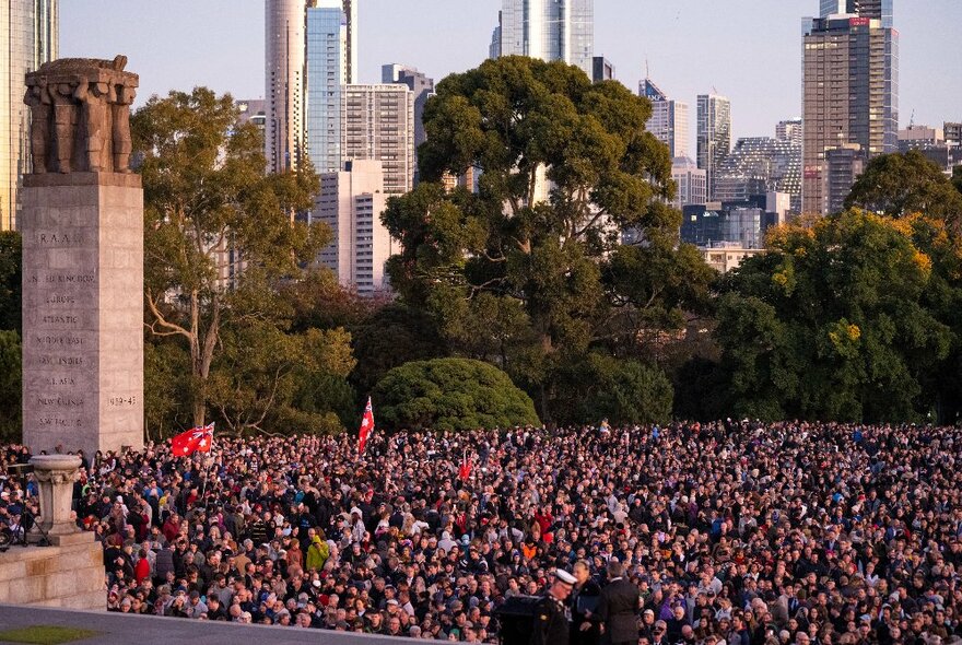 Large crowds of people gathered in the early morning on the forecourt at the Shrine of Remembrance, with Melbourne city building skyline and trees in the background.