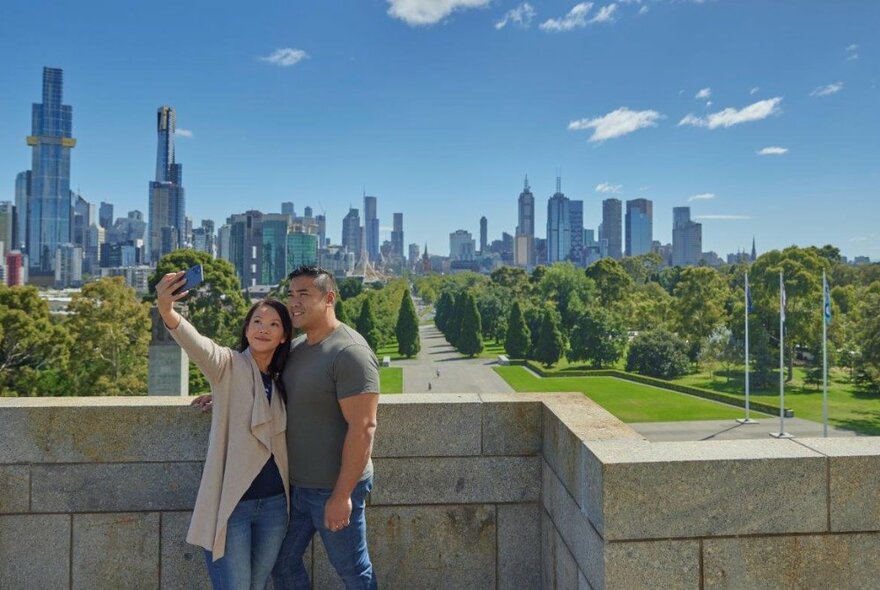 A couple taking a selfie at the Shrine of Remembrance with Melbourne's skyline behind.
