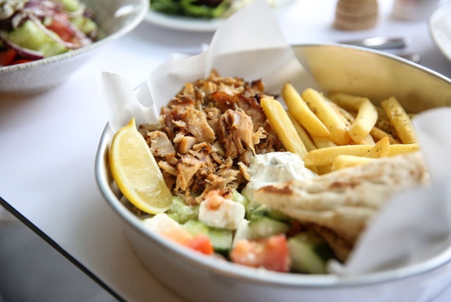 Looking at  metal bowl containing gyros, chips, pita and Greek salad with a slice of lemon. 