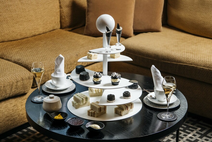 Afternoon tea setting on a round table beside chairs, with three-tiered cake and sandwich display, glasses of bubbles and dishes.