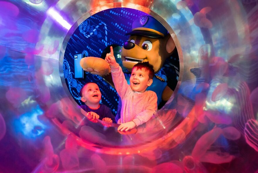 A person in a Chase costume from PAW Patrol with two young children looking through a circular window to colourful sealife beyond.