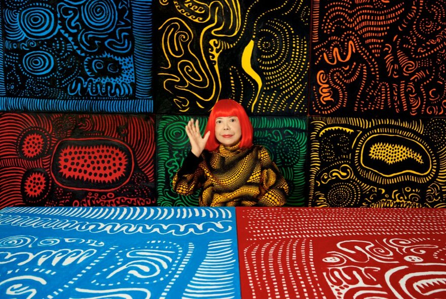 The artist Yayoi Kusama, with a bright red bob hairstyle, seated in front of a selection of her painted artworks displayed on a wall behind her and a table in front of her. 