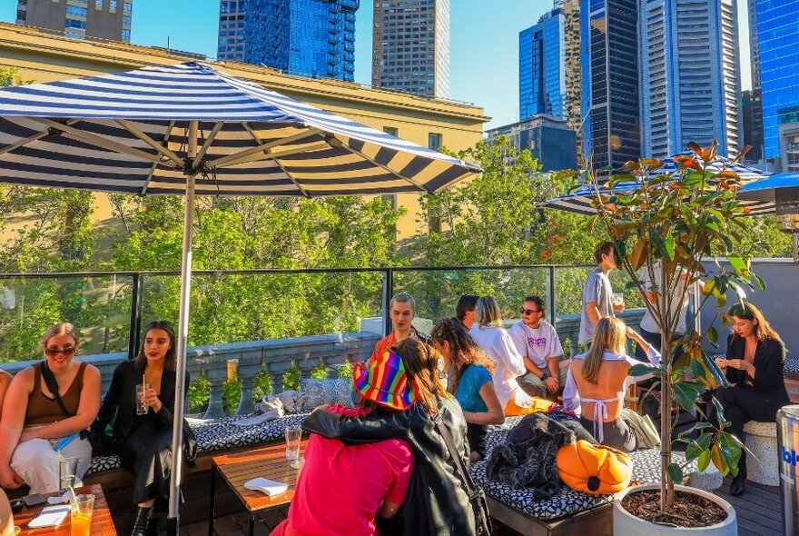 A busy rooftop bar with umbrellas and trees overlooking the city skyline.