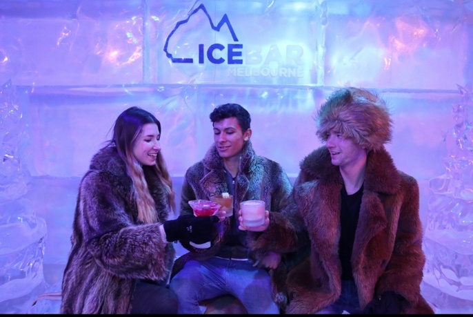 Three people wearing fur coats clinking glasses against an ice backdrop.