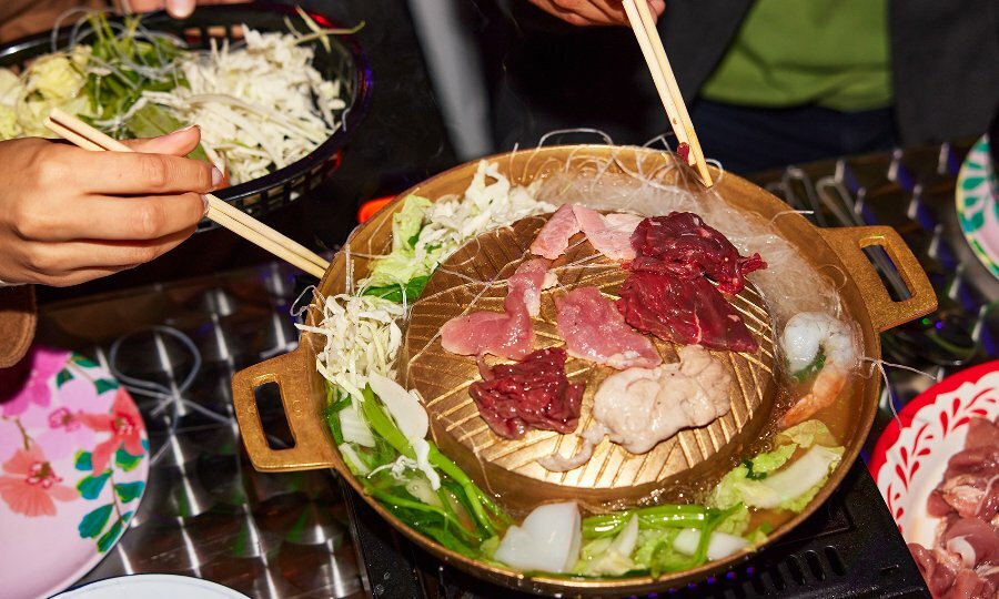 People cooking meat on a gold mookata hotpot grill with noodles and veggies in the broth.