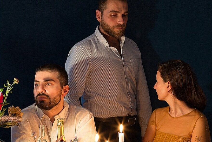 Three people at a candle-lit table; one man standing, another man seated facing to left, woman next to him turned around to face standing man.