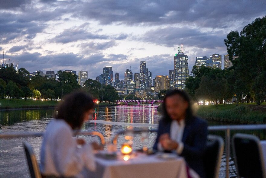 A blurred image of two people at a private table with a candle on the river with the city in the background. 
