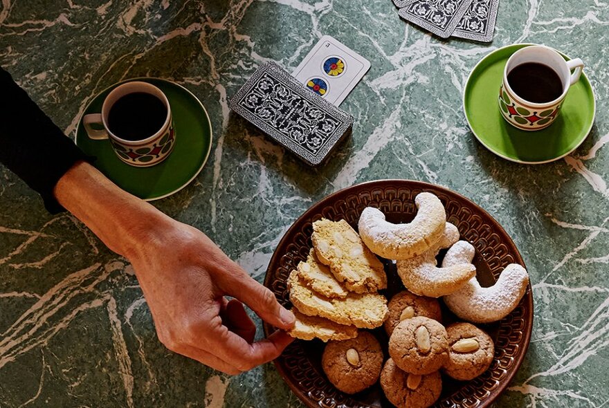 Looking down at a table with a hand selecting a biscuit from a plate, small coffee cups and a pack of Italian picture playing cards.