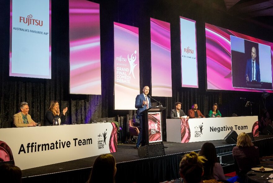 Person speaking at a lectern with two debating teams sitting at desks either side on a stage, vertical screens behind displaying company names.