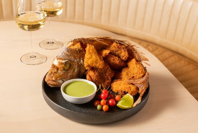 A whole fried fish with garnish and dipping sauce on a dark plate, on a table with two glasses of white wine alongside.