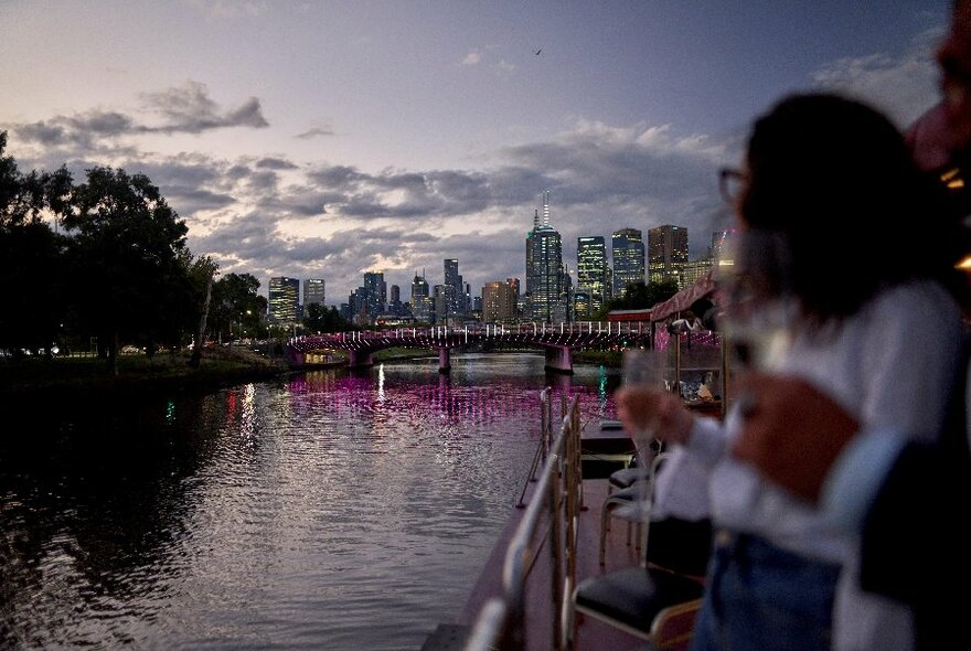 A blurred image of people on a boat with champagne flutes with the river and the city skyline in the background. 